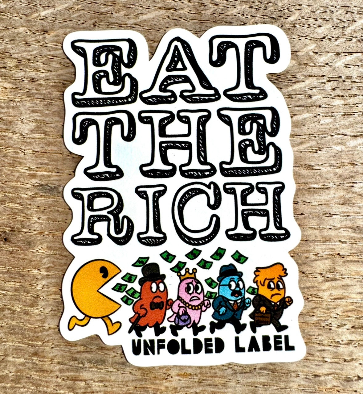 Protest sticker with 'Eat the Rich' text and cartoon characters on a wooden surface by Unfolded Label