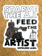 Sticker reads "Starve AI Feed the Artist" with robot graphic on a wooden surface by Unfolded Label